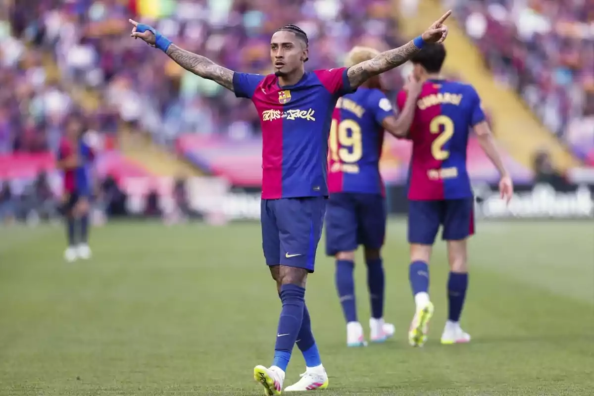 Un jugador de futbol amb uniforme blau i vermell aixeca els braços al camp mentre altres jugadors estan al fons. Un jugador de futbol amb uniforme blau i vermell aixeca els braços al camp mentre altres jugadors estan al fons.