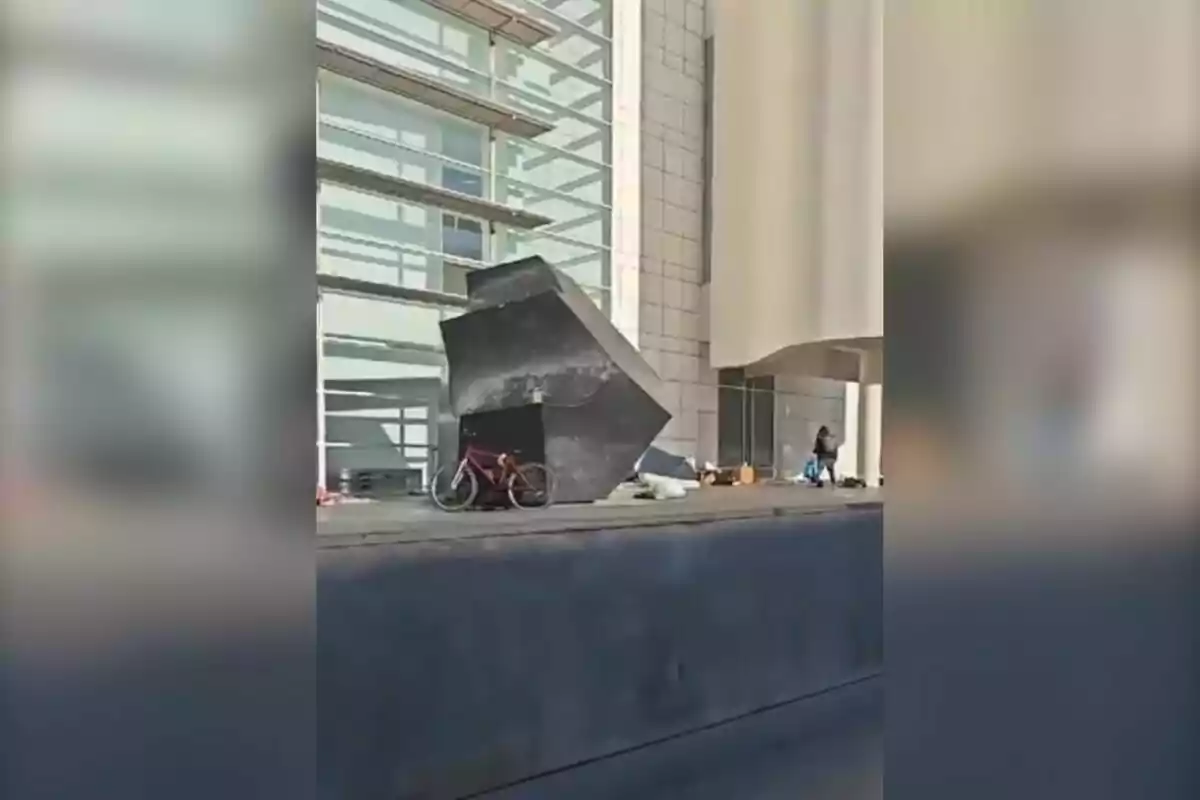 Una bicicleta vermella està estacionada al costat d´una escultura geomètrica negra davant d´un edifici modern amb parets de vidre i concret.