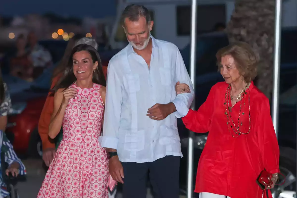 La reina Letícia, Felip VI i la reina Sofia somriuen mentre caminen junts a l’aire lliure a la nit, una dona amb vestit rosa, un home amb camisa clara i una dona gran amb brusa vermella. La reina Letícia, Felip VI i la reina Sofia somriuen mentre caminen junts a l’aire lliure a la nit, una dona amb vestit rosa, un home amb camisa clara i una dona gran amb brusa vermella.