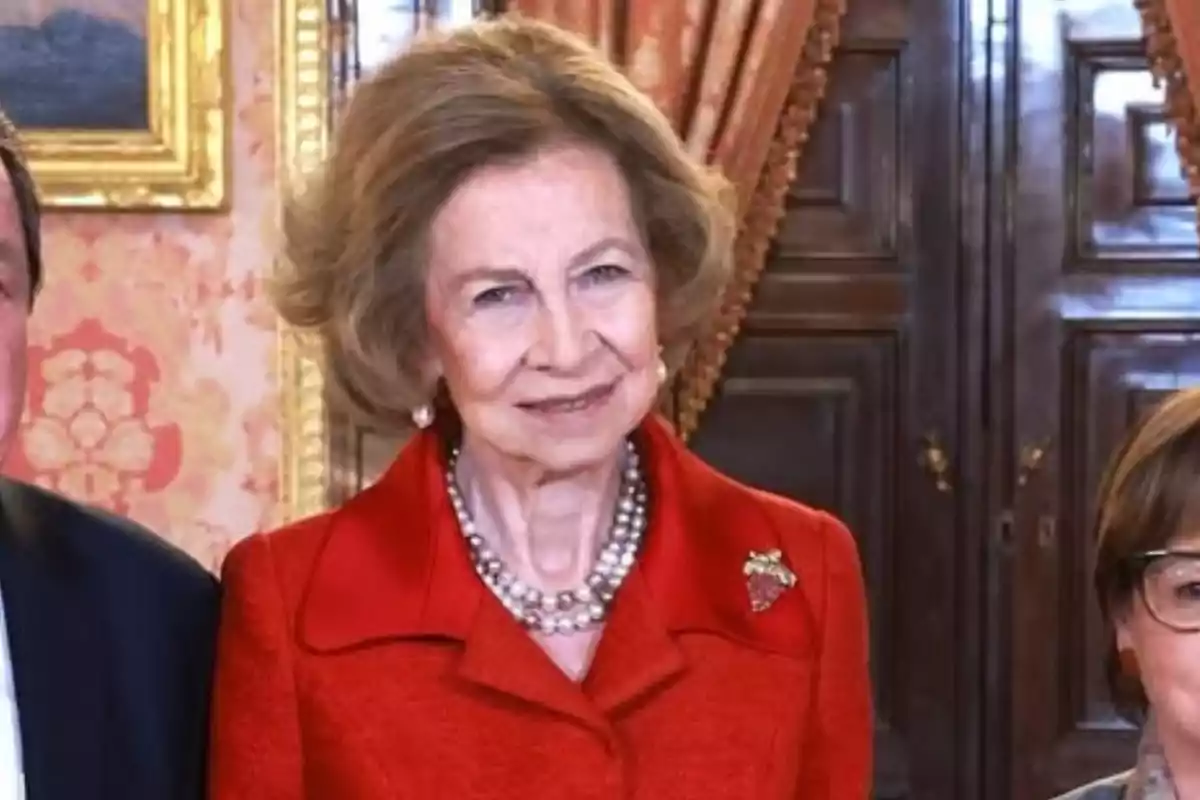 An older woman with light brown hair and a red suit smiles as she stands in an elegantly decorated room.