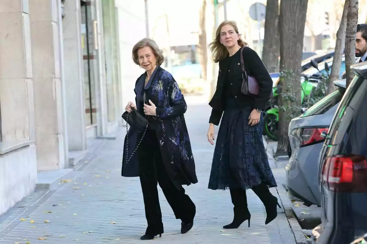 Queen Sofía and Infanta Cristina walking along a sidewalk next to a row of parked cars.