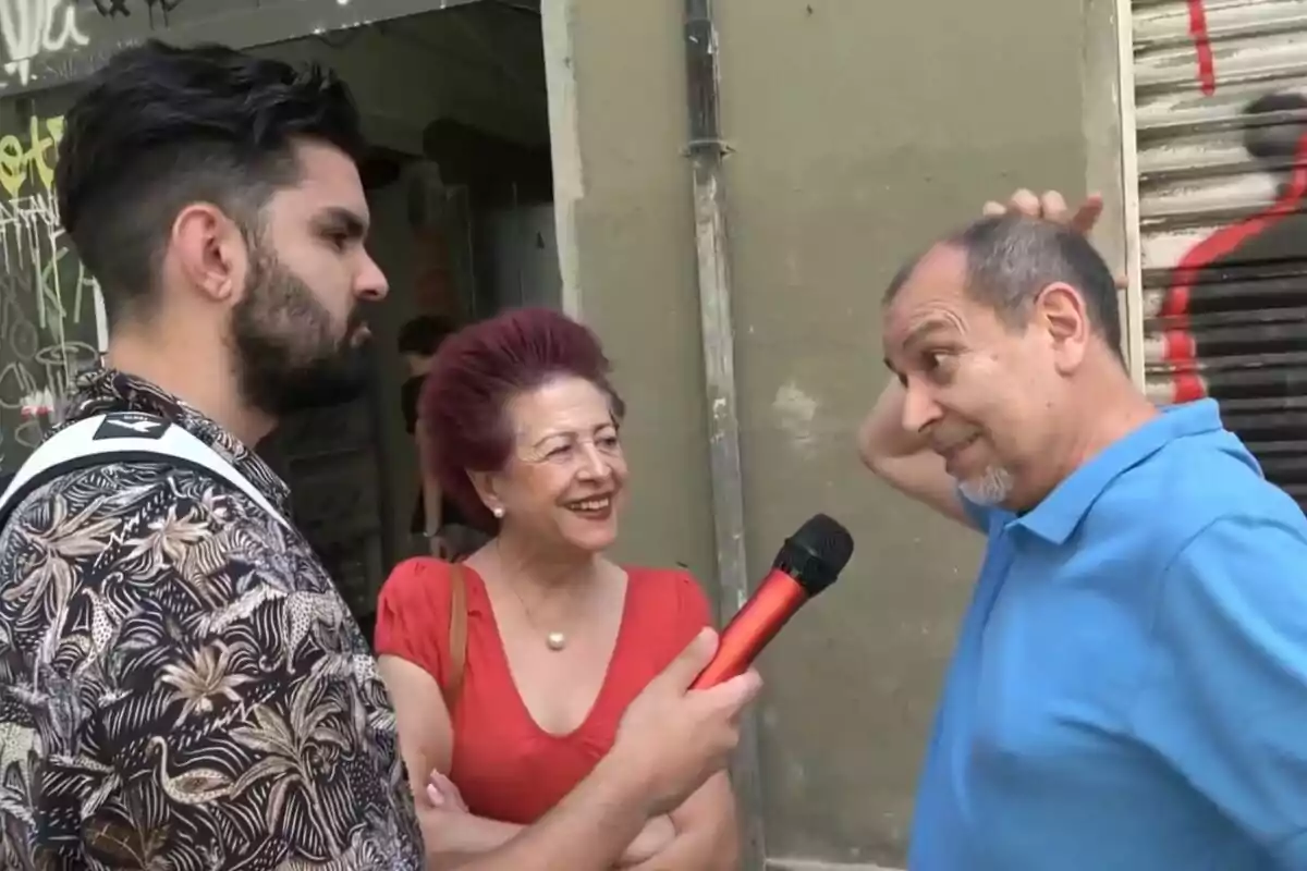 Un home amb barba sosté un micròfon vermell mentre entrevista una parella gran al carrer, la dona somriu i l?home es grata el cap.