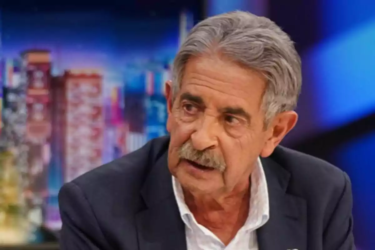 Miguel Ángel Revilla amb bigoti i cabell canós, vestit amb un vestit fosc i camisa blanca, en un entorn de fons acolorit i desenfocat a 'El Hormiguero'.