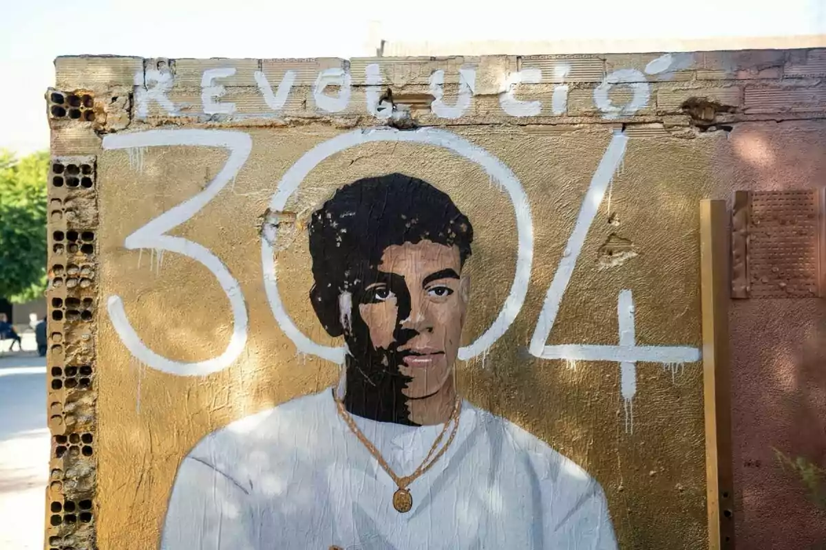 Un mural en una pared de ladrillos muestra el retrato de Lamine Yamal con cabello oscuro y una camiseta blanca, acompañado de las palabras "Revolució 304" pintadas en blanco sobre un fondo dorado.