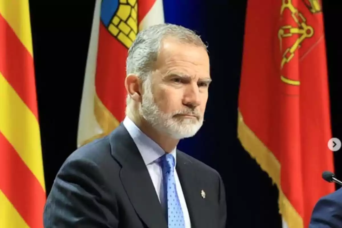 A bearded man in a dark suit stands in front of several colorful flags.