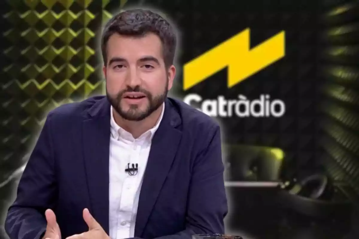 A man with a beard and a dark jacket is speaking in front of a background with a radio station's logo.