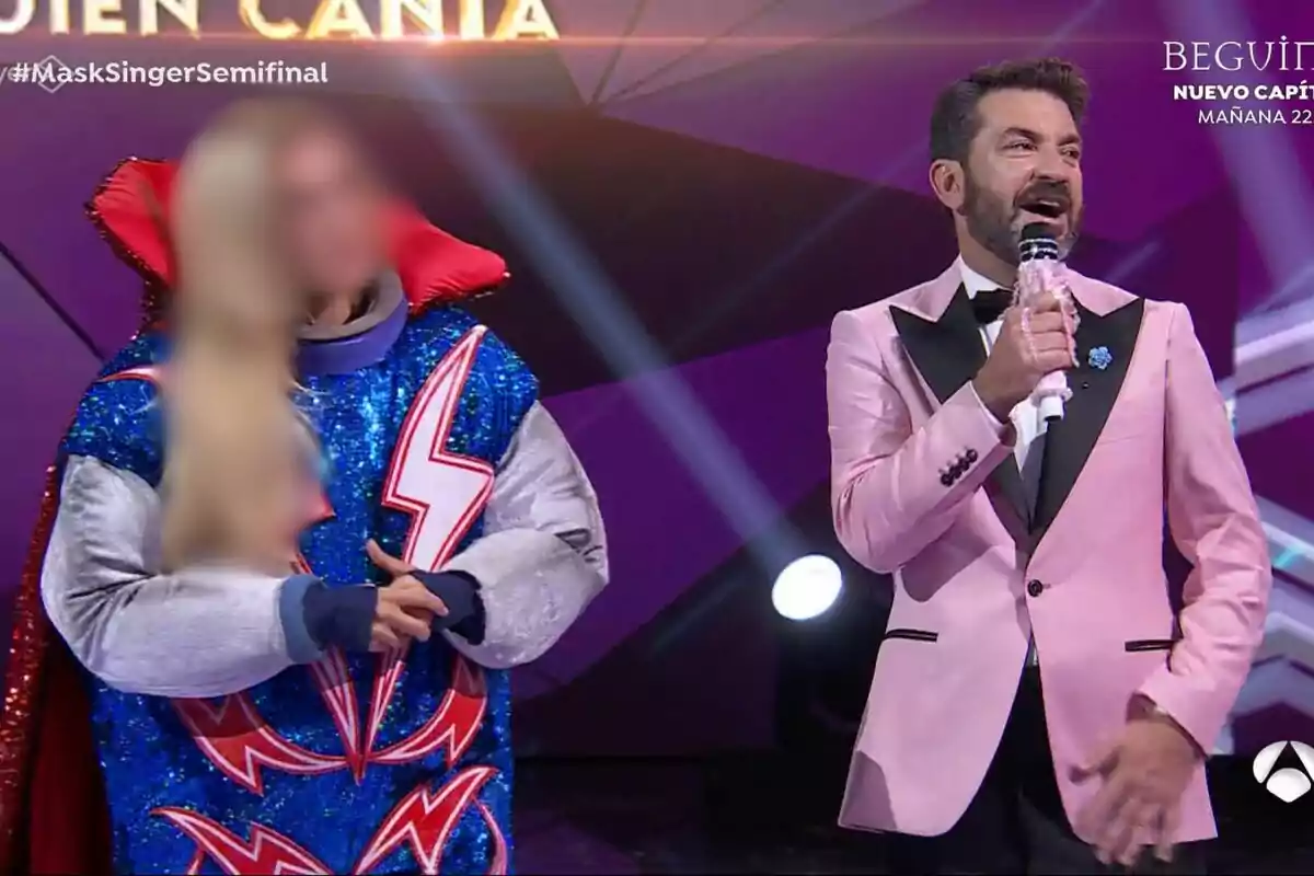 Arturo Valls amb un vestit rosa sosté un micròfon mentre està dret amb una persona disfressada amb un vestit blau brillant i detalls de raigs en un escenari il·luminat a Mask Singer.