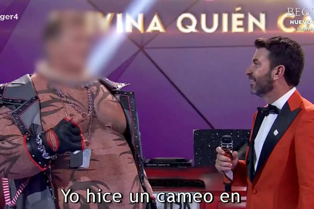 Arturo Valls amb un vestit taronja sosté un micròfon mentre parla amb David Hasselhoff amb la cara difuminada disfressat en un escenari de Mask Singer.