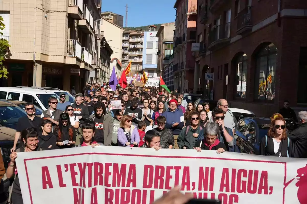 Un grup de persones participa en una manifestació en un carrer, sostenint una pancarta amb un missatge en català, envoltats d'edificis i sota un cel clar.