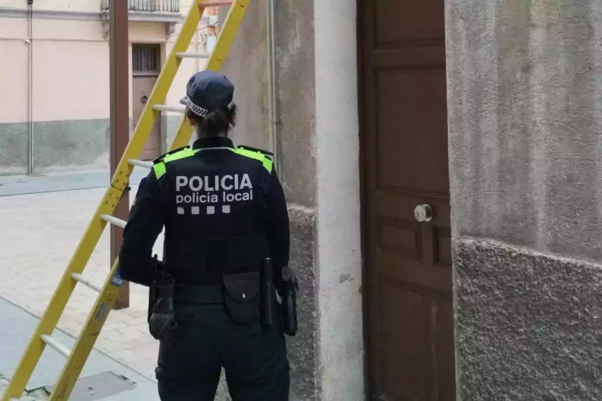 Una agente de la policía local de espaldas frente a una escalera amarilla apoyada en una pared junto a una puerta marrón.