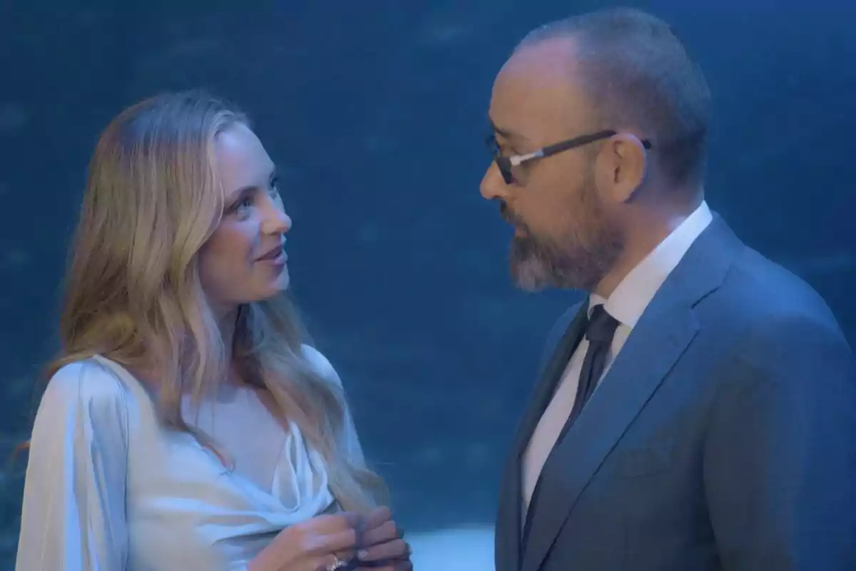 Laia Grassi i Risto Mejide conversen en un ambient il·luminat amb llum blava, ella porta un vestit clar i ell vesteix un vestit formal amb ulleres a Viajando con Chester.