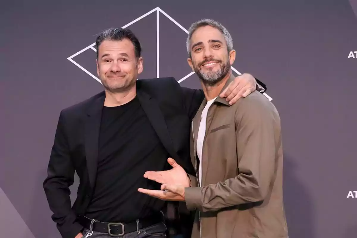 Iñaki López i Roberto Leal posant junts i somrient davant d'un fons gris amb un disseny geomètric blanc a la presentació de López i Leal contra el canal.