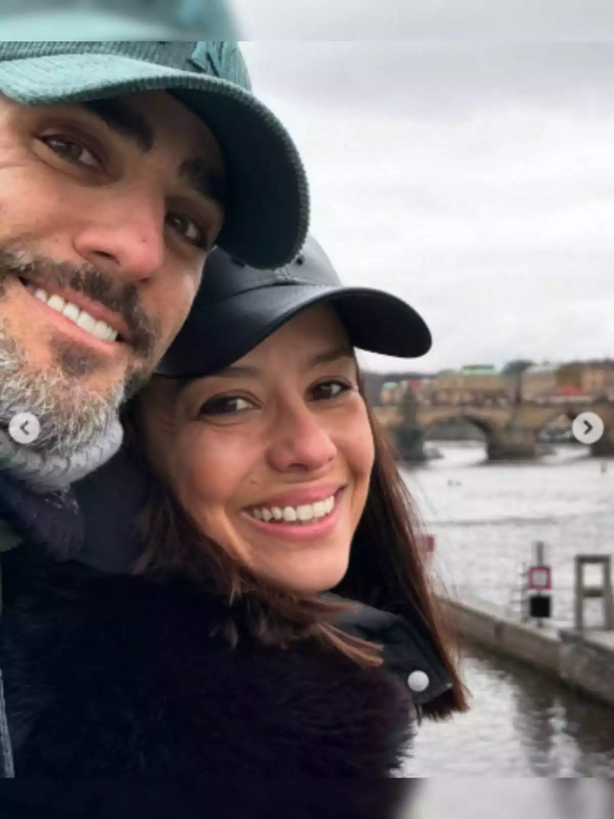 A smiling couple—composed of Sara Rubio and Roberto Leal—wearing caps posing in front of a river and a bridge on a cloudy day. A smiling couple—composed of Sara Rubio and Roberto Leal—wearing caps posing in front of a river and a bridge on a cloudy day.