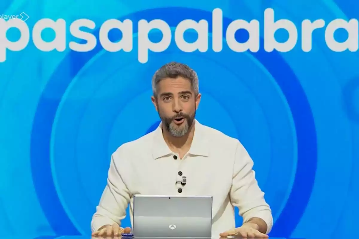 Roberto Leal está frente a una pantalla azul con el logo de 'Pasapalabra' detrás de él, usando una camisa blanca y hablando al público.