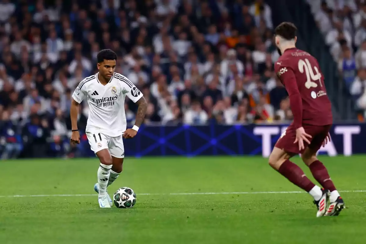 A player in a white uniform controls the ball as an opponent in a brown uniform approaches in a stadium full of spectators.