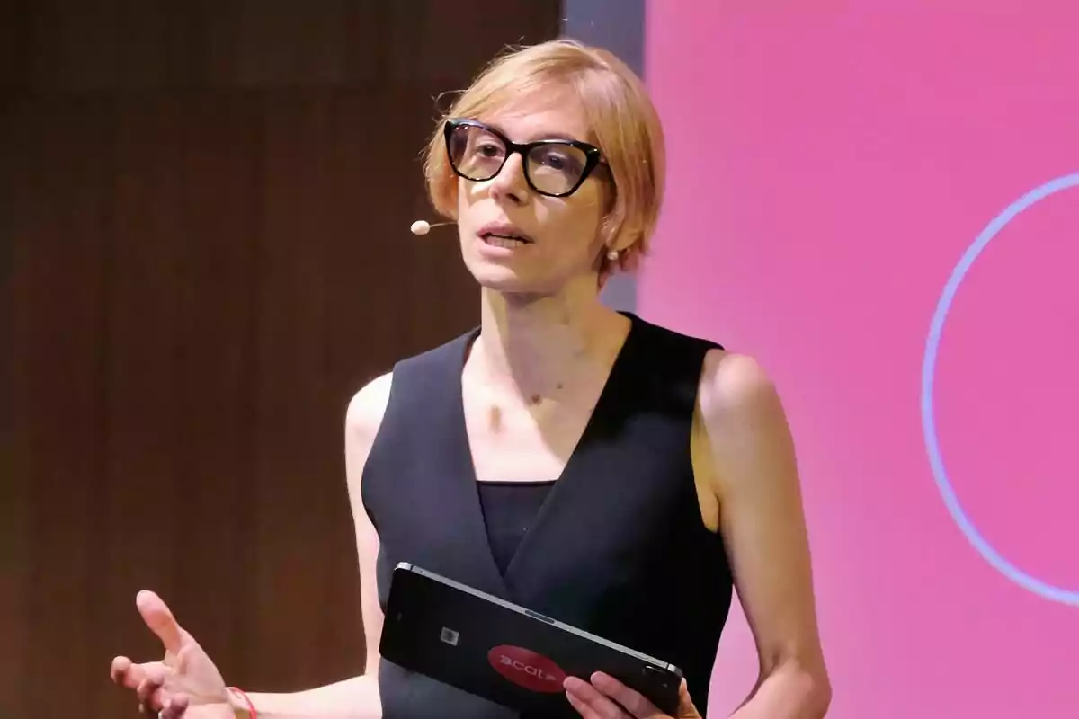 Rosa Romà de cabell curt i ros amb ulleres grans i vestit negre parlant en públic mentre sosté una tauleta davant d’un fons rosa de 3Cat