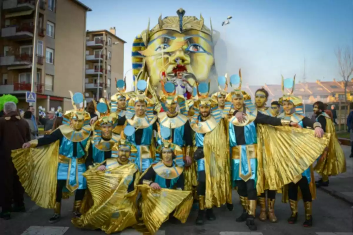 Un grupo de personas disfrazadas con trajes inspirados en el antiguo Egipto posan frente a una gran figura de una cabeza faraónica en un entorno urbano.