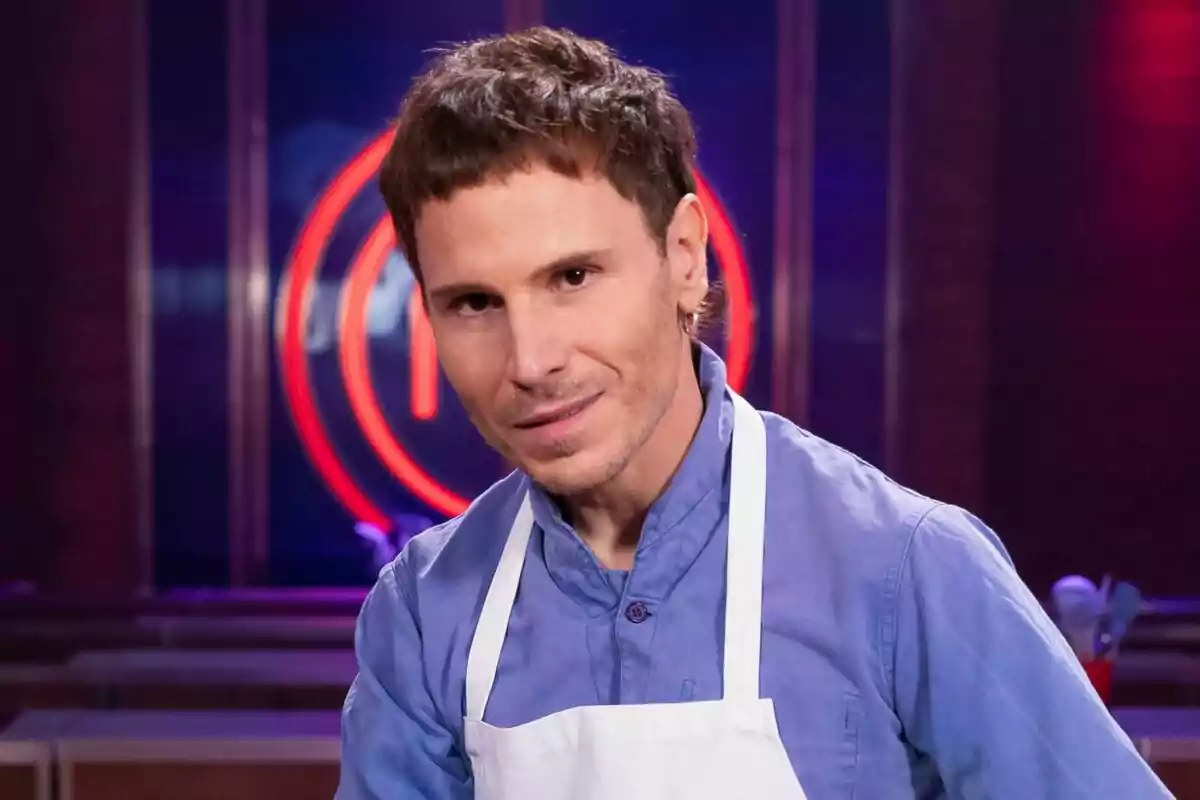 Rubén Ochandiano amb davantal blanc i camisa blava en un set de cuina amb un logotip vermell de MasterChef Celebrity al fons.