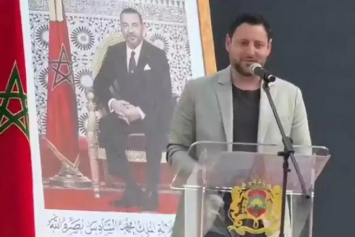 Un home parlant en un faristol amb un micròfon, al costat d'un retrat d'una figura oficial i una bandera amb un símbol d'estrella.