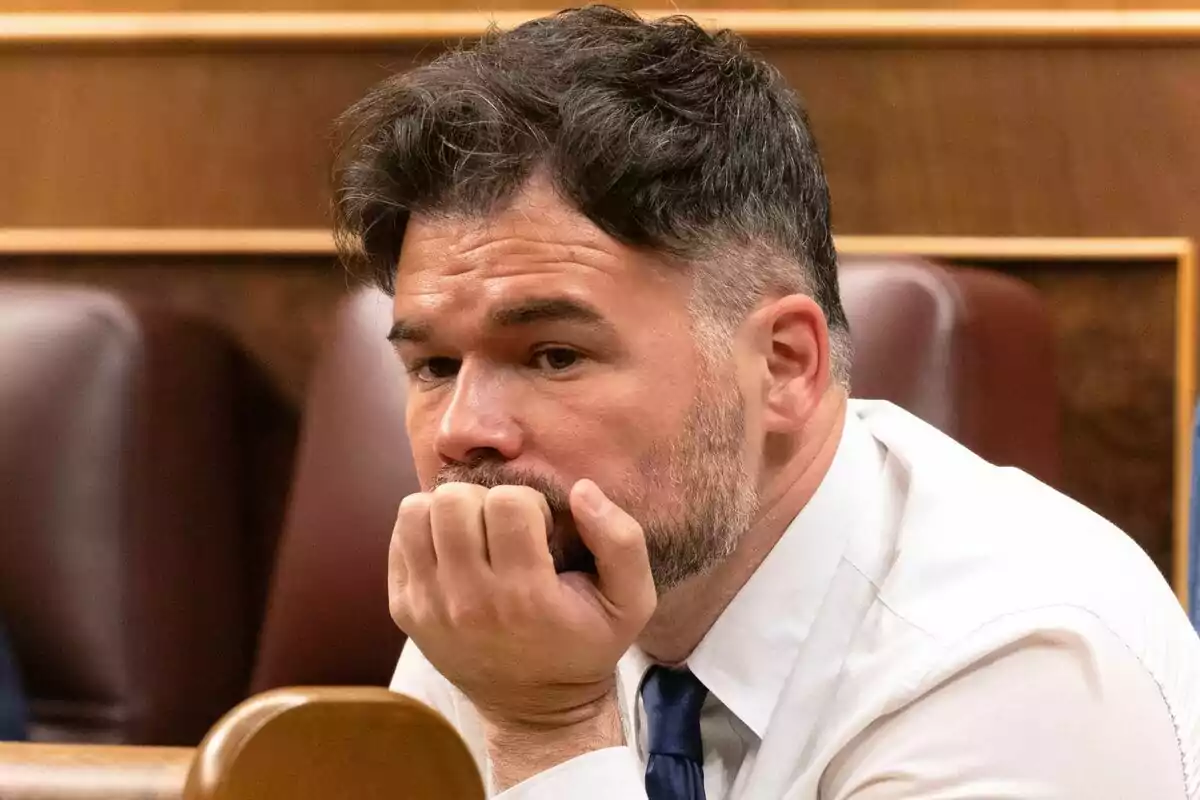 Gabriel Rufián amb camisa blanca i corbata blava assegut en una sala, amb la mà a la barbeta i expressió pensativa.