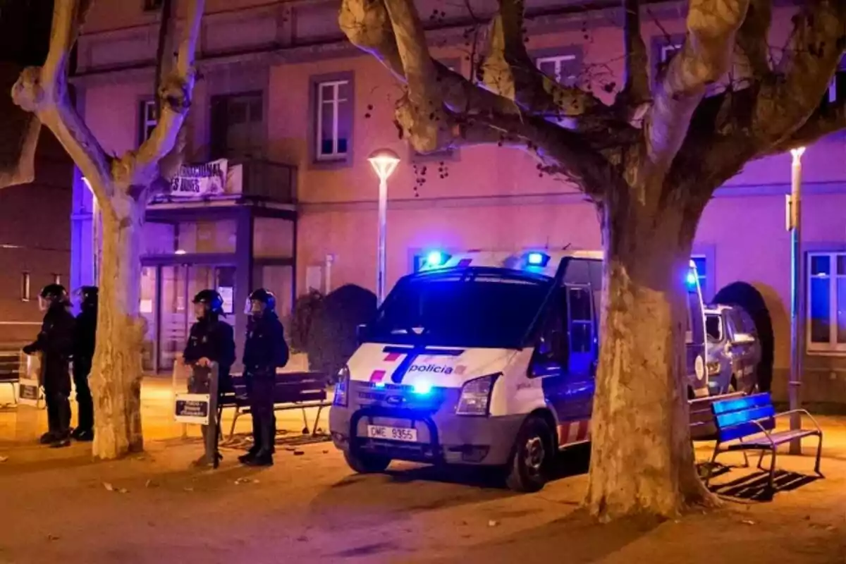 Un grup de policies amb equip antidisturbis està dret al costat d'una furgoneta policial amb llums blaves enceses en un carrer arbrat durant la nit.