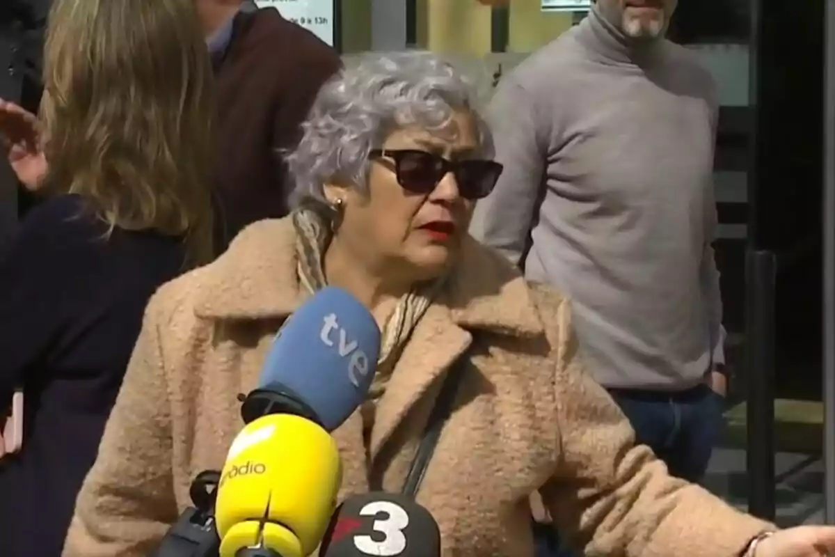 Una mujer con gafas de sol y abrigo claro está frente a varios micrófonos de diferentes medios de comunicación mientras otras personas están en el fondo.