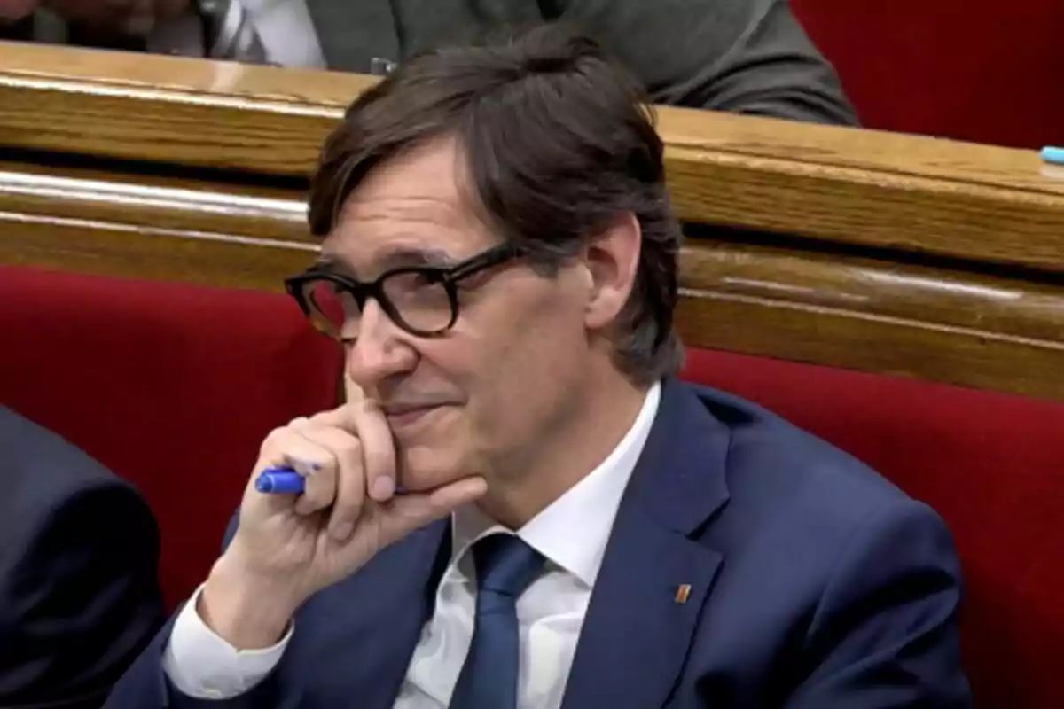 Un home amb ulleres i vestit blau sosté un bolígraf blau mentre està assegut en un seient vermell.
