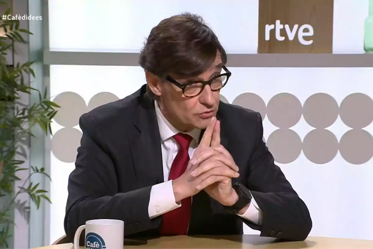 Un home amb ulleres i vestit fosc està assegut a una taula, amb les mans entrellaçades, davant d'una tassa que diu "Cafè" i un fons amb el logotip de rtve.