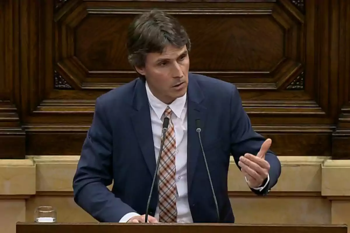 Un home en vestit parlant en un faristol amb fons de fusta.