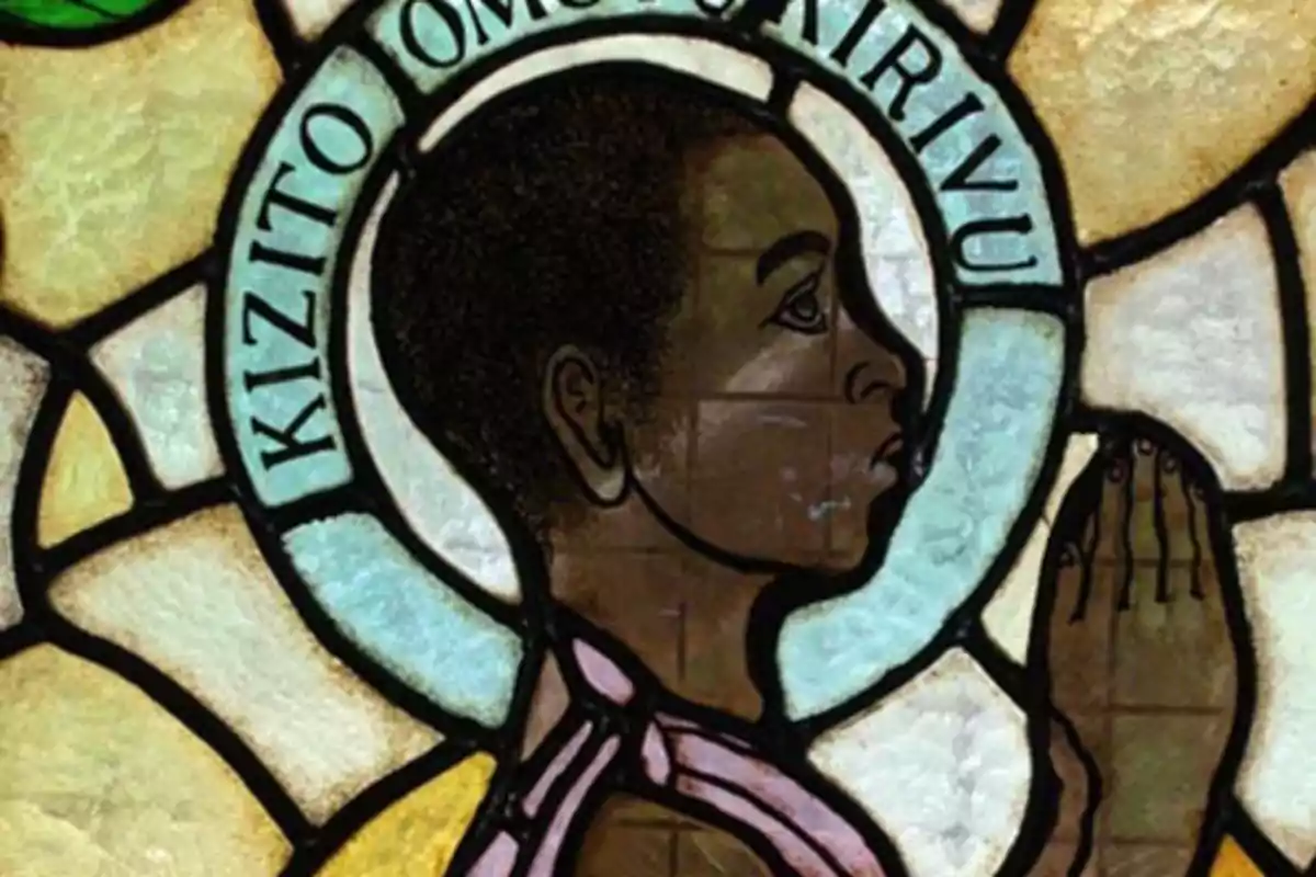 Vitrall amb la imatge d’un jove de perfil resant amb les mans juntes i un halo blau al voltant del cap on es llegeix el nom Kizito