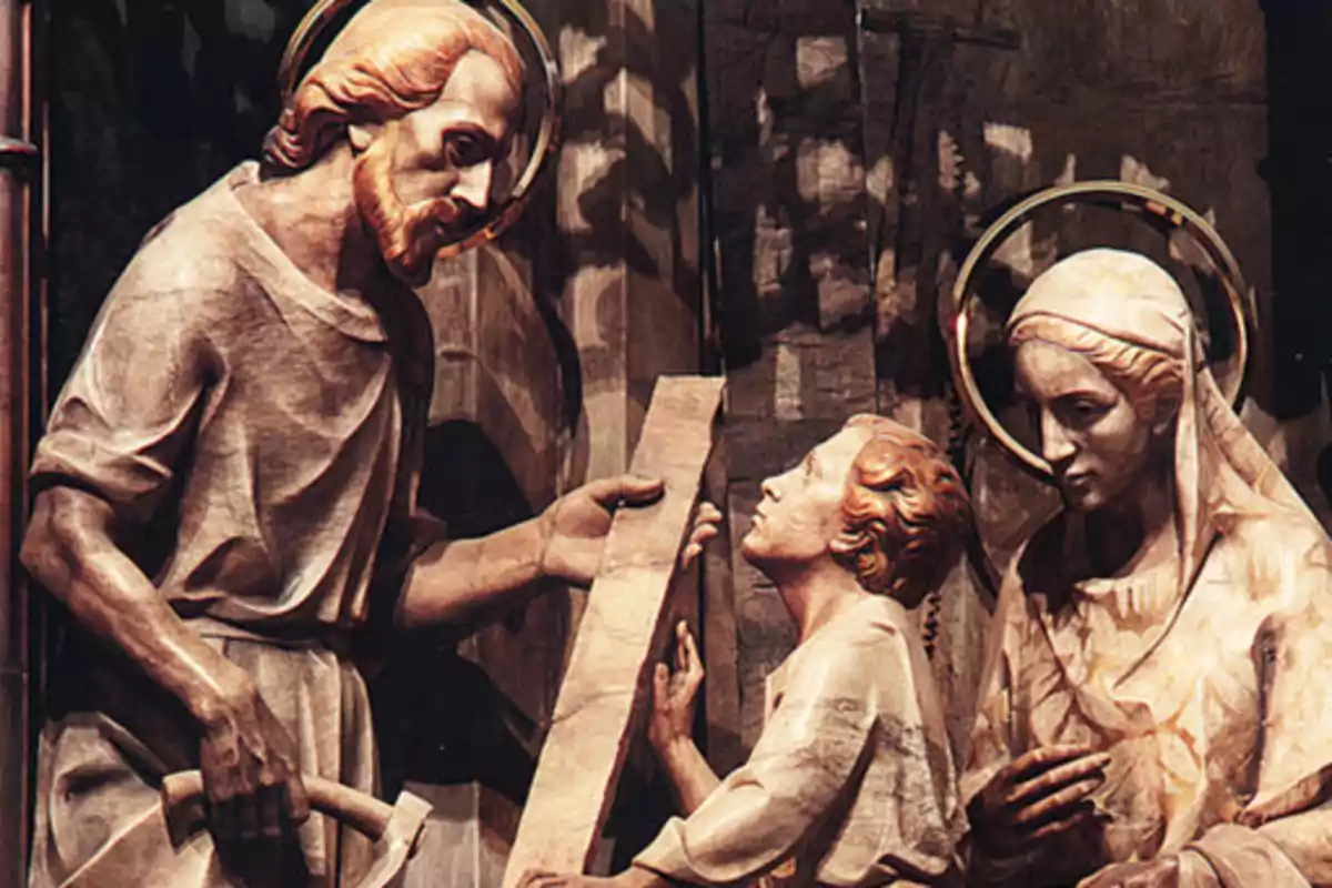 Escultura de tres figures religioses en una escena d'ensenyament, amb un home sostenint una eina i una taula, un nen observant i una dona mirant amb atenció.