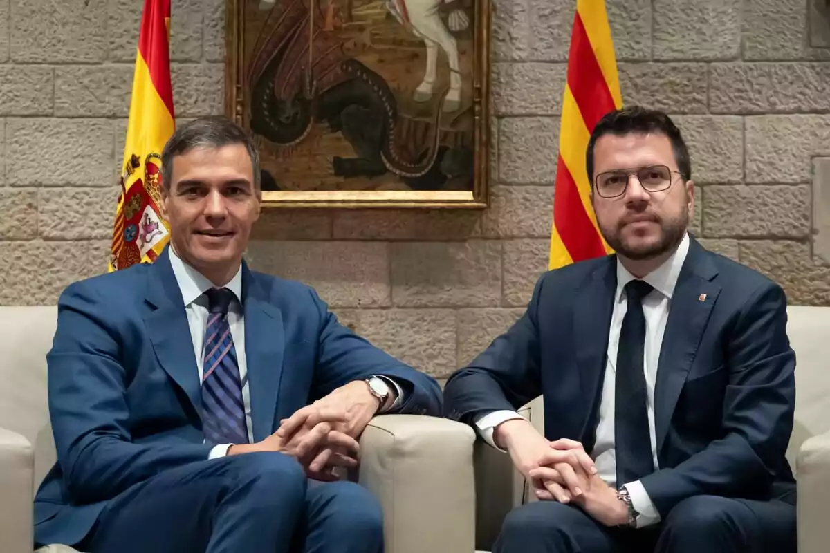 Dos homes amb vestits formals asseguts en butaques, amb banderes d'Espanya i Catalunya darrere seu i un quadre a la paret.