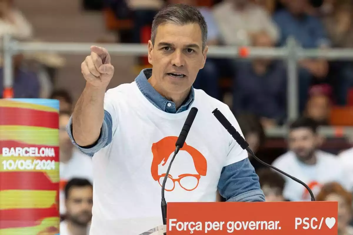 Pedro Sánchez en un míting parlant des d'un faristol amb una samarreta amb el símbol de la cara de salvador illa i assenyalant al capdavant