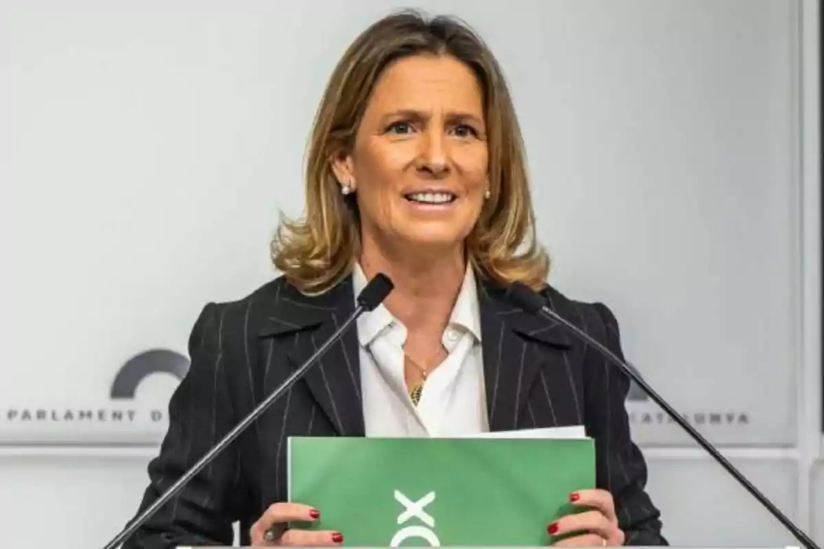 Blonde, short-haired woman speaking at a press conference with two microphones and holding a green folder