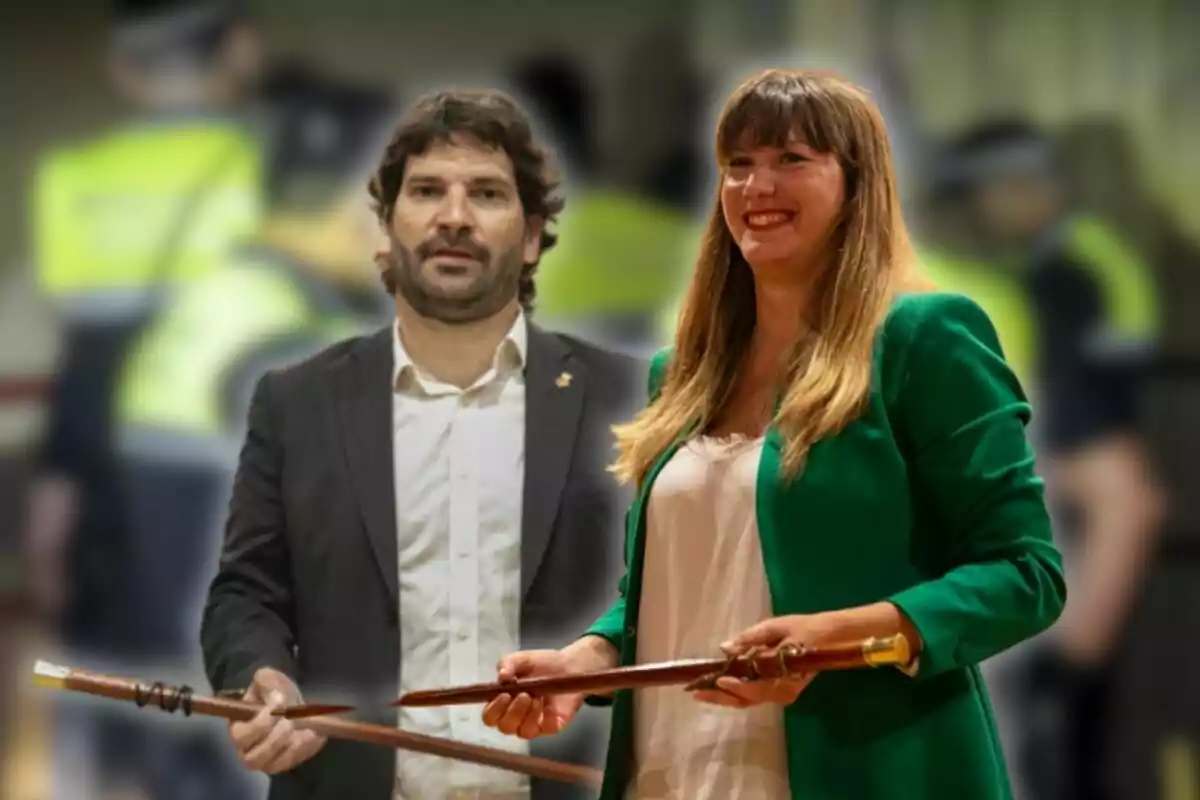 Dues persones dretes, una dona amb jaqueta verda i un home amb vestit fosc, tots dos sostenint bastons, amb un fons desenfocat de persones amb armilles grogues.