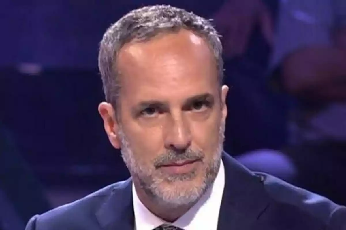 Santi Acosta con barba y cabello corto canoso viste traje oscuro y mira hacia adelante en un entorno iluminado en De Viernes.