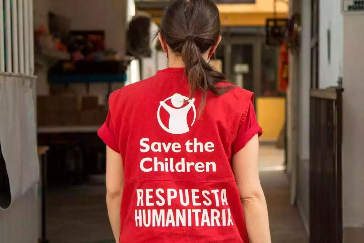 Pla mitjà d'una voluntària de l'ONG Save the Children d'esquena i amb una armilla vermella amb el logo de l'ONG i la frase 'resposta humanitària'