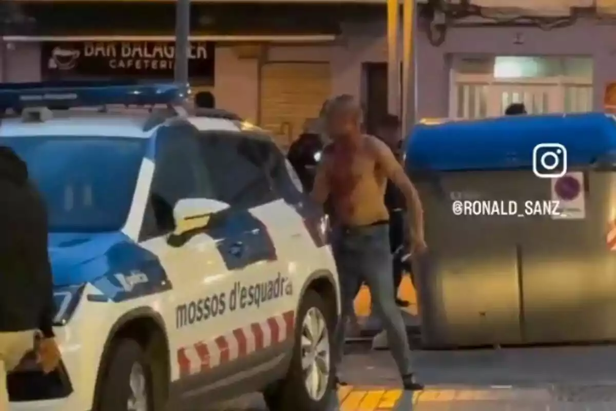 Un home sense camisa està dret al costat d'un cotxe de policia dels Mossos d'Esquadra en un carrer, amb un contenidor d'escombraries blau al fons i una cafeteria visible darrere.