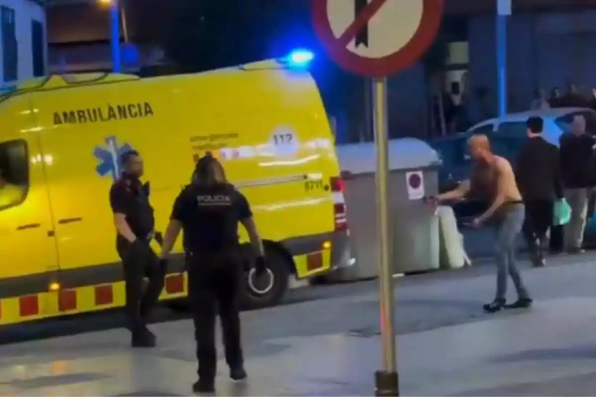 Una ambulància groga està estacionada al carrer mentre dos policies interactuen amb un home sense camisa a prop d'un contenidor d'escombraries. Una ambulància groga està estacionada al carrer mentre dos policies interactuen amb un home sense camisa a prop d'un contenidor d'escombraries.