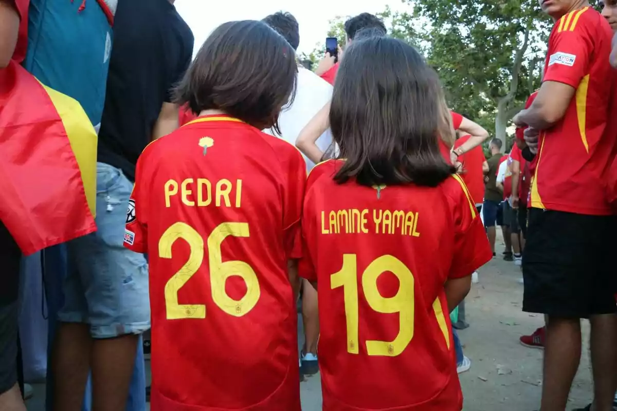 Dos nens d'esquena porten samarretes vermelles de la selecció espanyola de futbol amb els noms "Pedri" i "Lamine Yamal" i els números 26 i 19, envoltats de més persones amb samarretes similars.