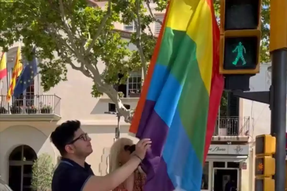 Una persona sostenint una bandera de l'orgull LGBT+ amb un semàfor en verd per a vianants en un carrer amb arbres i edificis de fons.