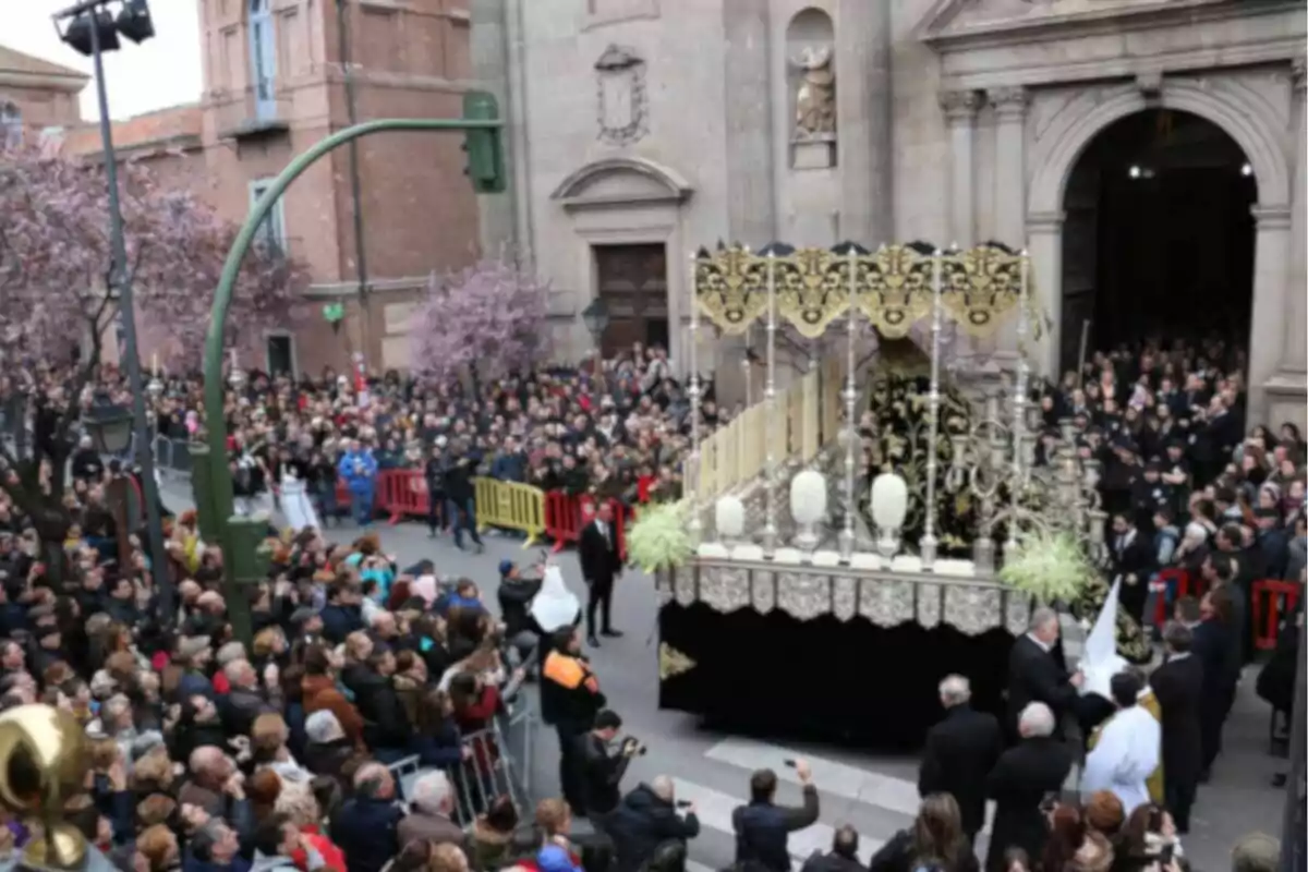 Una multitud se congrega en una calle para observar una procesión religiosa con un paso adornado con detalles dorados y plateados frente a una iglesia.