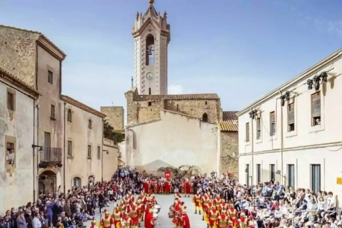 Un desfile de personas vestidas con trajes rojos y amarillos en una plaza rodeada de edificios antiguos con una torre de reloj al fondo y una multitud observando.