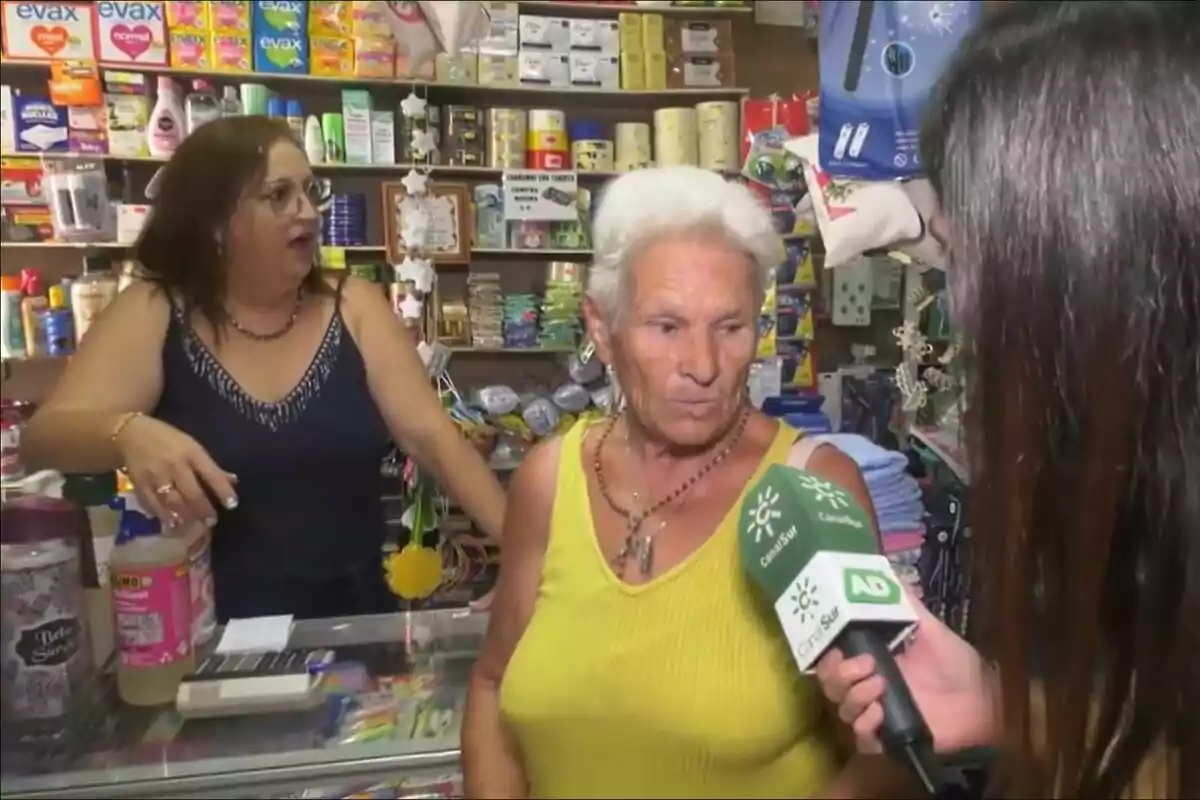 Una dona gran amb cabell blanc i samarreta groga està sent entrevistada en una botiga per a Andalusia Directe a Canal Sur, mentre una altra dona amb ulleres i samarreta fosca observa des del taulell ple de productes.