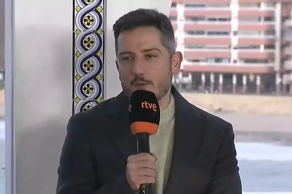 Un hombre con barba sostiene un micrófono de RTVE mientras está de pie frente a un fondo urbano y decoraciones de azulejos en Benidorm Fest.