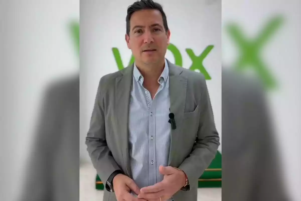 Un hombre de pie con chaqueta gris y camisa azul claro frente a un fondo con el logotipo de Vox.