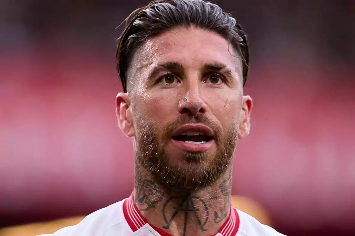 A bearded soccer player with tattoos on his neck wears a white jersey with red details. A bearded soccer player with tattoos on his neck wears a white jersey with red details.