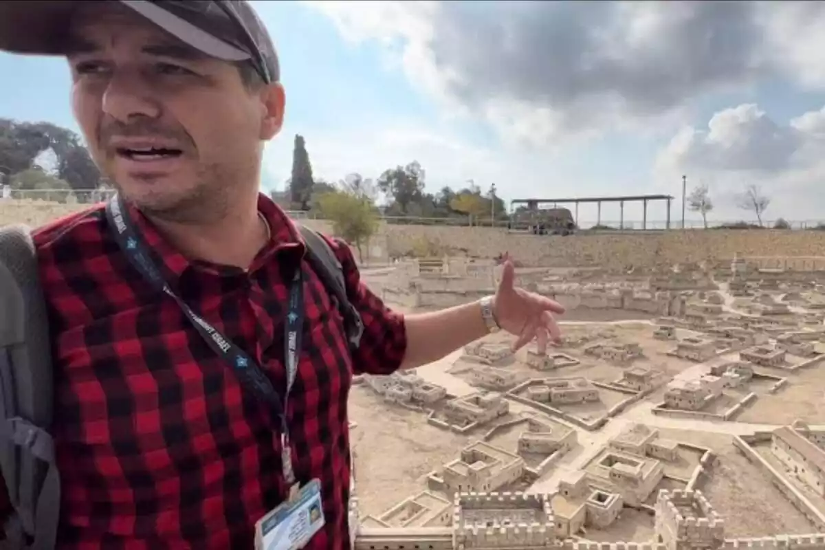 Pla mitjà de Shimon Edry amb camisa de quadres i gorra parlant sobre unes restes arqueològiques que apareixen al fons de la imatge