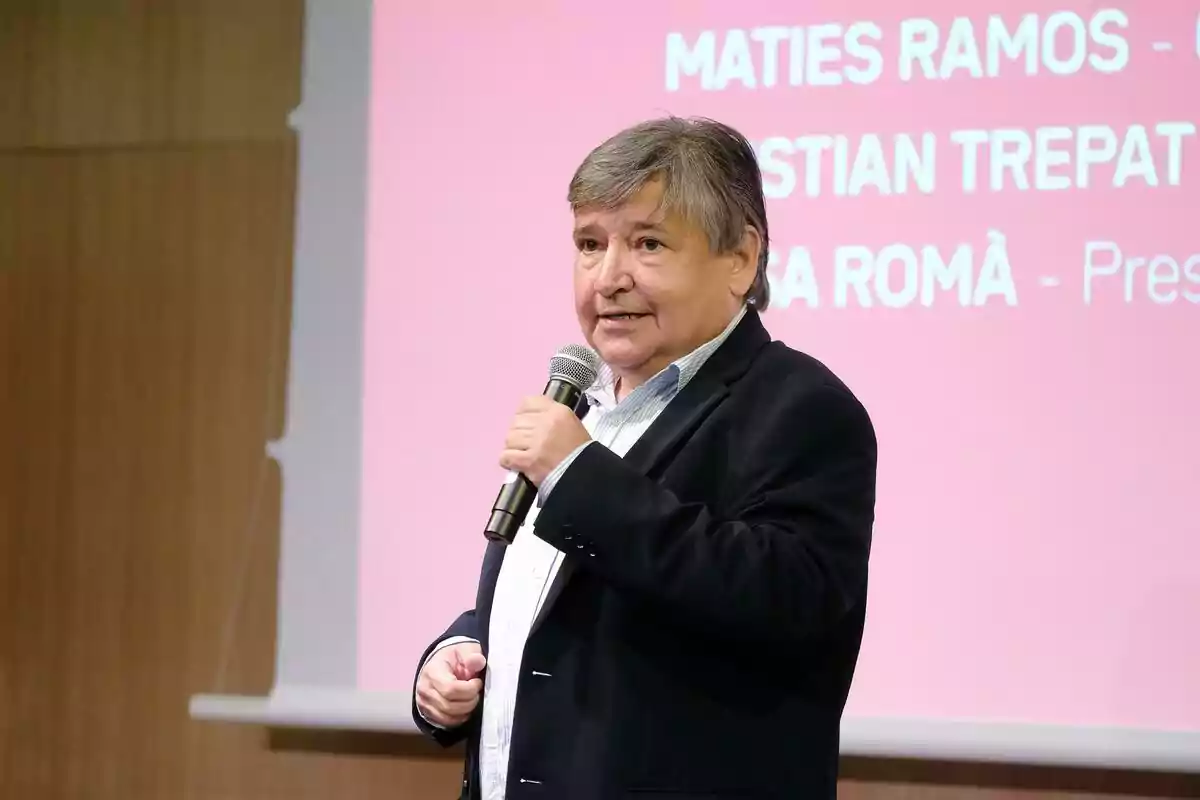 Sigfrid Gras de cabells canosos parlant per micròfon davant d’una pantalla amb fons rosa i text blanc de 3Cat. Sigfrid Gras de cabells canosos parlant per micròfon davant d’una pantalla amb fons rosa i text blanc de 3Cat.