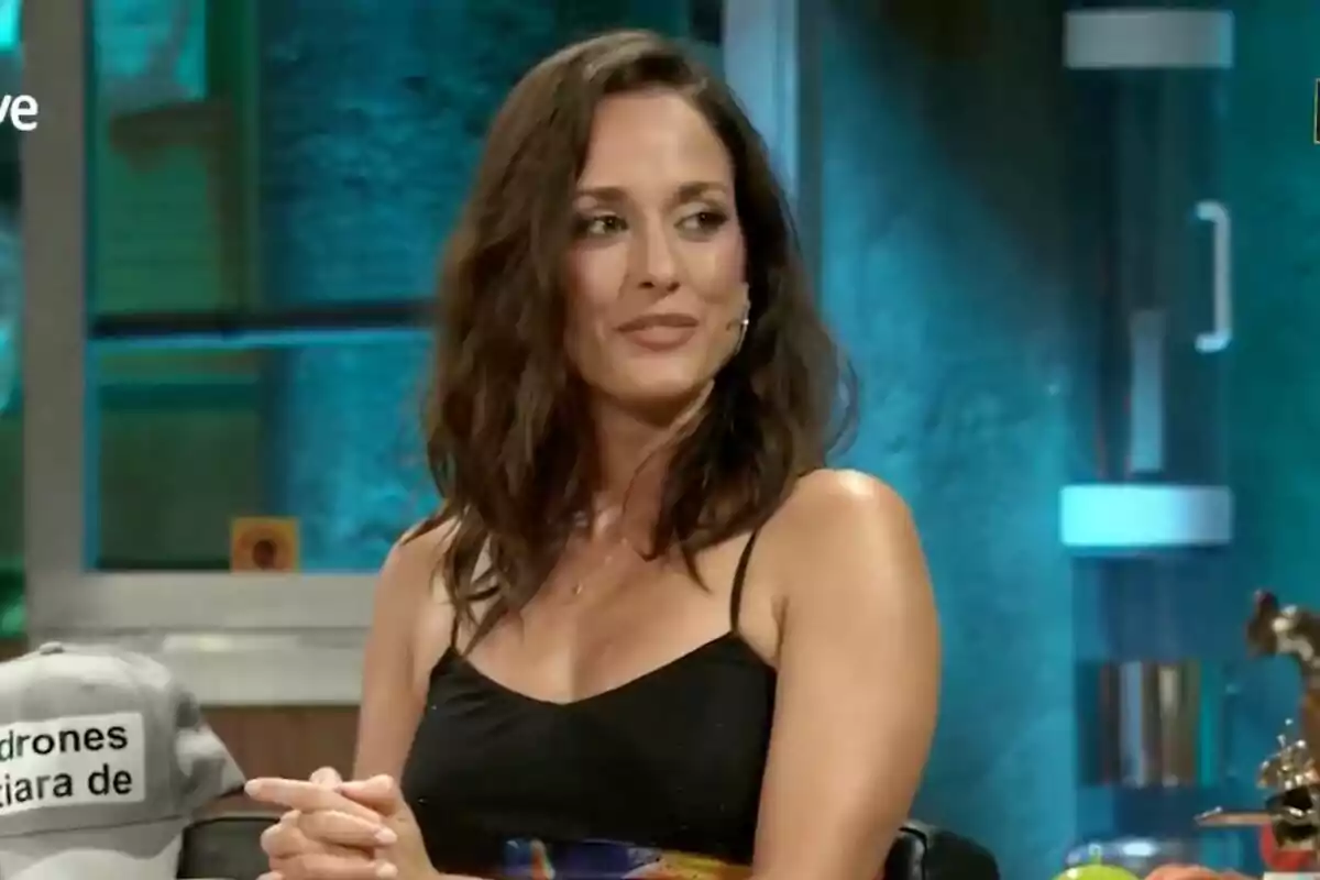 Sílvia Alonso amb cabell castany i ondulat asseguda a 'La Revuelta' amb fons blau i una gorra grisa al seu costat